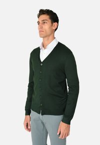 Cardigan verde scuro, tessuto a maglia, design a scollatura a V, sei bottoni, polsini e orlo a costine, indossato sopra una camicia bianca con colletto, stile aderente.