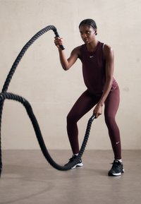 Bordó sportos ujjatlan felső és leggings, fekete edzőcipő, fekete harci kötelek fogása egy zárt térben. Simább textúrák, formás kivitel.