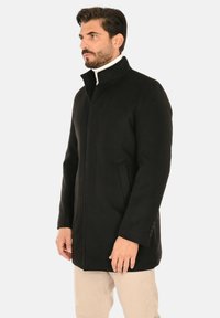 Cappotto nero con collo alto, caratterizzato da un taglio dritto e tasche laterali. Tessuto liscio con design minimalista e senza chiusure visibili.