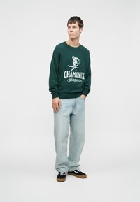 Zeleni pleteni pulover z napisom "CHAMONIX Francija" in grafiko smučarja. Ujemajoče lahke modre kavbojke in črne superge s svetlim podplatom.