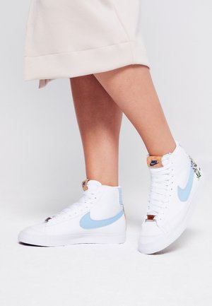 Henkilö, jolla on valkoiset Nike-tarrakengät vaaleansinellä Swoosh-logolla ja kukkakuviolla kantapäässä, yhdistettynä kermanväriseen hameeseen.