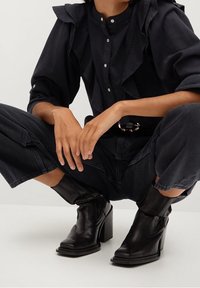 Botas de tobillo de cuero negro con tacones cuadrados, que presentan detalles de costura y un acabado ligeramente texturizado, usadas con un atuendo amplio y oscuro.