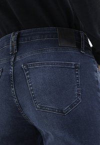 Dunkelblaue Denim-Jeans mit einer glatten Textur, ausgestattet mit einem schwarzen Lederpatch am Bund und hinteren Taschen mit dezenten Steppnähten.