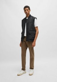 Zwarte mouwloze vest met een knoopsluiting aan de voorkant en een borstzak, gecombineerd met een wit T-shirt, bruine broek en witte sneakers.