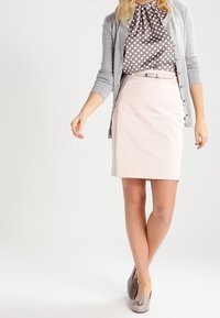 Kvinde iført grå polkaprikket bluse, lys grå cardigan, bleg rosa knyttet nederdel og taupe kvast loafers, stående med benene krydset.
