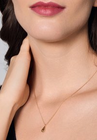 Złoty naszyjnik z łańcuszka z wiszącym pendantem w kształcie kropli, gładką powierzchnią i błyszczącym wykończeniem, spoczywający na gładkiej skórze w okolicy obojczyka.