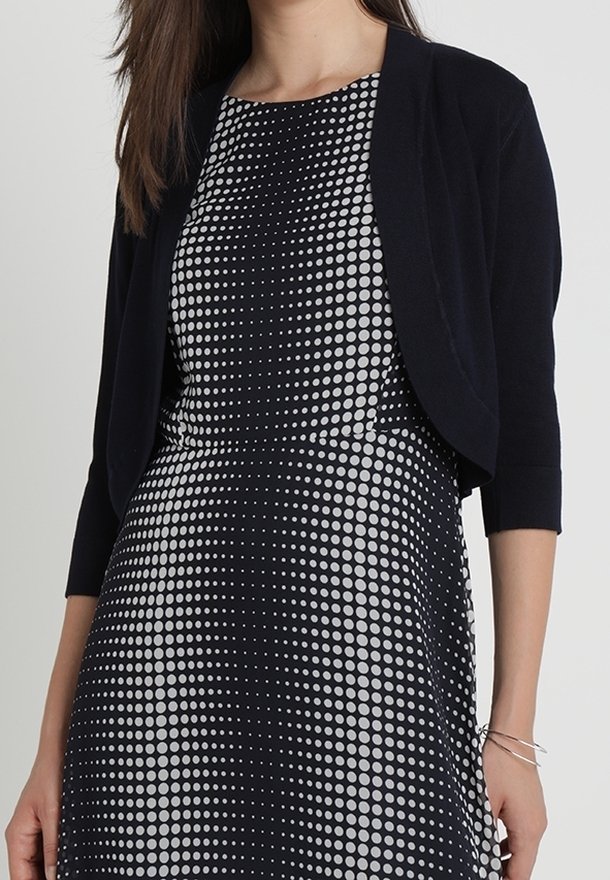 Laivastonsininen bolerotakki avoimella edestä, mustavalkoisen pilkkumekon päällä, siinä on vartalonmyötäinen leikkaus ja kolme neljänneksen mittaiset hihat.