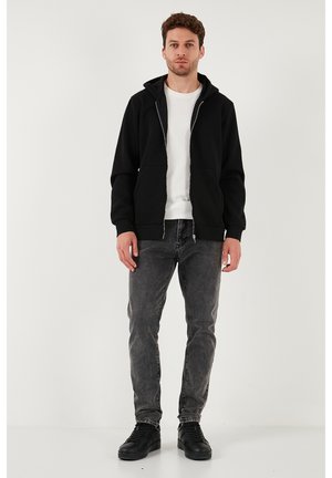 Schwarzer Hoodie mit Reißverschluss über einem weißen Shirt, kombiniert mit grauen Jeans und schwarzen Sneakers. Der Hoodie hat eine vordere Tasche und eine Kapuze mit Kordelzug.