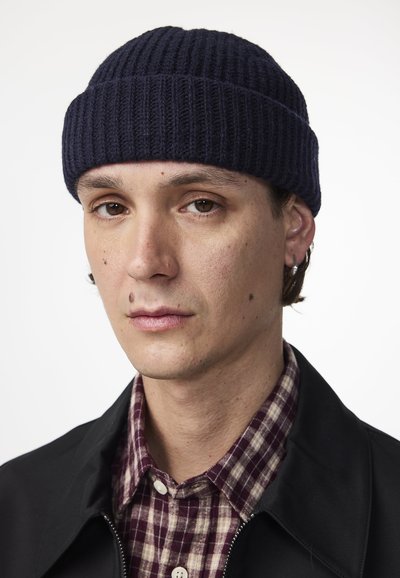 Granatowa dziana czapka typu beanie z prążkowanym wzorem, noszona z czarną kurtką i bordowo-kremową koszulą w kratę. Skóra pokazuje kilka piegów.