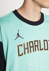 Camiseta verde menta con cuello negro. Presenta "CHARLOTTE" en letras negritas de color negro y dorado con un logo de Jordan negro arriba. Textura suave.