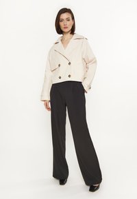 Licht beige dubbelzijdige jas met grote knopen en een brede kraag, gecombineerd met zwarte wijde broek en zwarte schoenen.