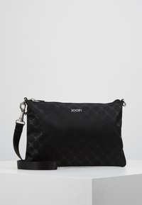 Sac bandoulière JOOP! noir avec motif subtil, fermoir argenté et bandoulière réglable, présenté sur une plateforme blanche sur fond gris.