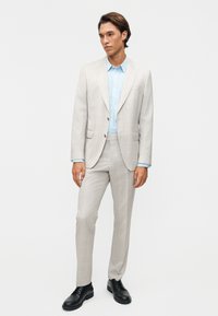 Άνδρας με ανοιχτό γκρι καρό κοστούμι, ανοιχτό μπλε πουκάμισο και μαύρα παπούτσια, στέκεται μπροστά σε απλό λευκό φόντο.