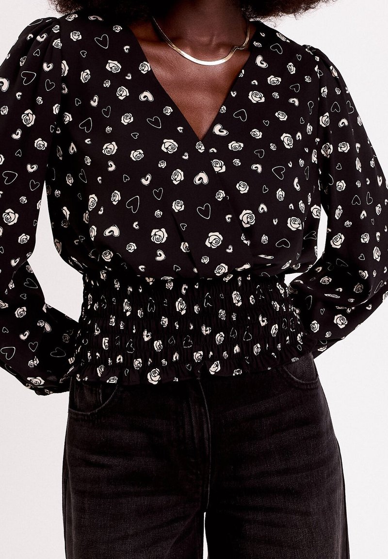 Femme portant un chemisier noir avec des motifs de roses blanches et de cœurs, manches bouffantes, taille smockée et pantalon noir taille haute.