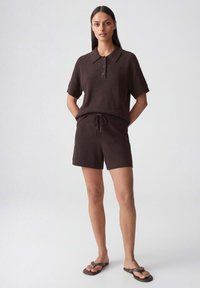 Woman standing wearing dark brown short-sleeve knit polo shirt, matching drawstring shorts, and dark brown flip-flops on plain background.
