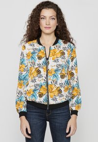 Giacca bomber floreale con sfondo bianco, presenta motivi di ibisco giallo e blu, colletto e polsini a coste neri, chiusura con zip.