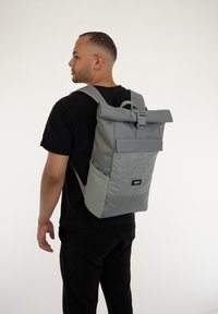 Sac à dos gris à enrouler avec un tissu texturé, des bretelles ajustables et une poche avant. Doté d'une fermeture à boucle et d'une étiquette de marque.