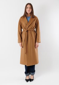 Cappotto marrone con cintura e un ampio colletto, realizzato in tessuto morbido. Presenta maniche lunghe, tasche laterali ed è abbinato a jeans scuri e tacchi neri.