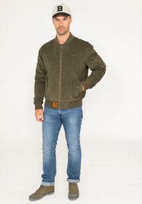 Veste bomber vert olive avec une texture lisse, des poignets côtelés et une fermeture éclair. Associée à un jean bleu clair et des bottes brunes.