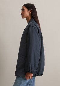 Donker marineblauwe pufferjack met een relaxte pasvorm, voorzien van een rits, geribbelde kraag en zijzakken, gecombineerd met lichtblauwe jeans.