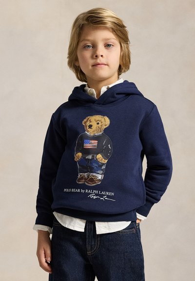 Bambino piccolo con capelli biondi che indossa una felpa blu navy con cappuccio raffigurante un orso con jeans e un maglione con la bandiera americana, con le mani nelle tasche.
