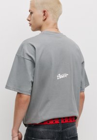 Lys grå oversized t-shirt med korte ærmer, der har et hvidt broderet logo på ryggen; båret med et rødt prikket bælte.