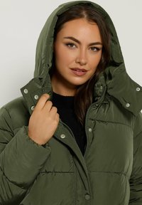 Mørkegrøn pufferjakke med hætte, der har en frontlynlås, trykknapper og quiltet tekstur. Hånden placeret nær kraven.