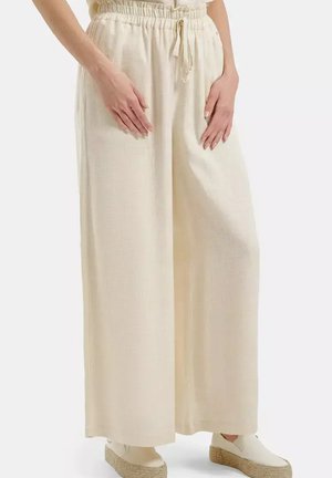 Kobieta nosząca luźne kremowe spodnie z wysokim stanem i sznurkiem w talii oraz białe buty na klinie typu slip-on, stoi z rękami spoczywającymi na udach.
