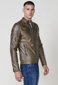 Uomo con una giacca di pelle marrone con tasche con zip e jeans blu, in piedi contro uno sfondo chiaro e semplice.