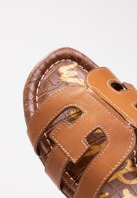 Sándalo de cuero marrón con un patrón de cocodrilo texturizado y diseños recortados. Presenta correas color crema y un logo dorado en contraste en la planta del pie.
