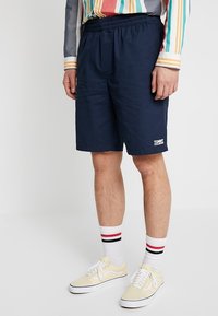 Námořnické modré krátké kalhoty s elastickým pasem, s malým bílým logem na lemu. Osoba má na sobě žluté boty a pruhované ponožky.