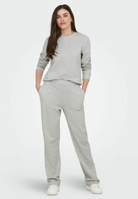 Vrouw met lang bruin haar, gekleed in een lichtgrijze sweater en bijpassende joggingbroek met witte sneakers, staande met handen in de zakken.