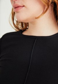 Nahaufnahme des unteren Gesichts einer Person, des Halses und des oberen Brustbereichs, die ein schwarzes geripptes Oberteil mit einer zentralen Naht trägt.