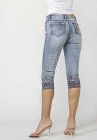 Woman wearing light blue cropped jeans with embroidered hems and clear high-heeled sandals, standing against a plain white background.