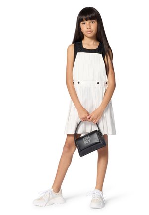 Ragazza con lunghi capelli scuri indossa un abito bianco a righe sottili sopra una maglia nera, scarpe da ginnastica bianche e tiene in mano una borsa nera DKNY.