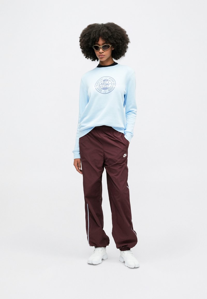 Sweatshirt bleu clair avec un logo circulaire, associé à un pantalon de survêtement bourgogne foncé avec des bandes blanches sur les côtés. Des baskets blanches complètent l'ensemble.