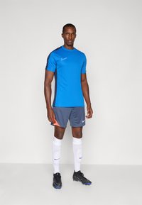 Sininen urheilupaita, jossa on tummansinisiä sivupaneeleja, harmaat shortit oranssilla korostuksella, valkoiset sukat ja mustat kengät, joissa jokaisessa osassa on Nike-logo.