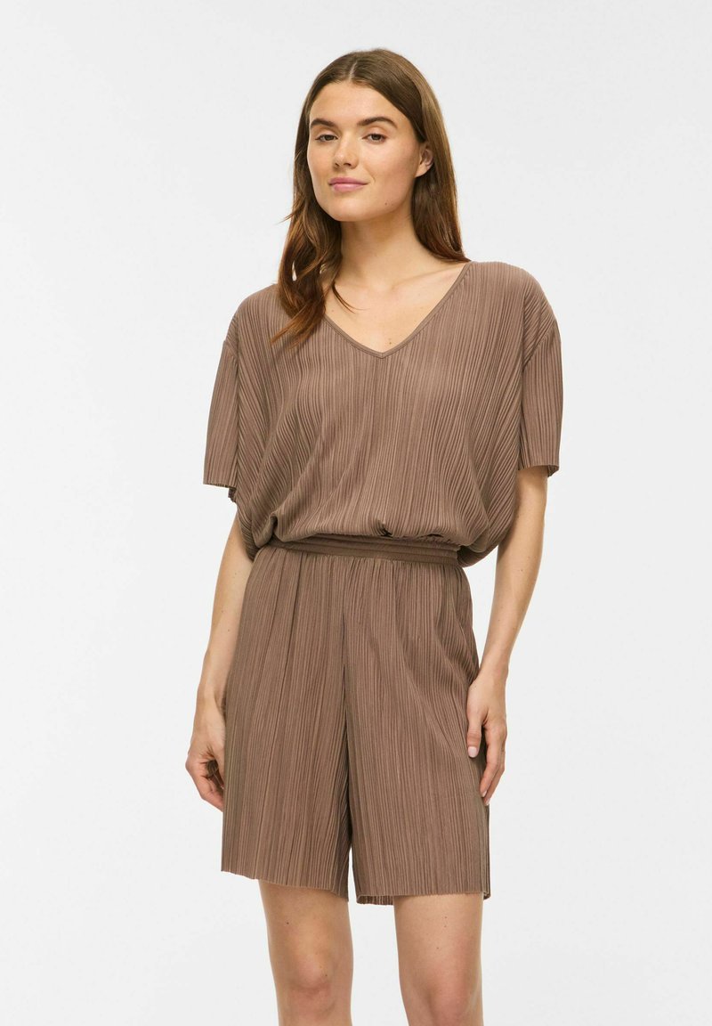 Jeune femme aux longs cheveux bruns portant un haut marron ample à manches courtes plissé et un short plissé assorti, debout devant un fond uni.