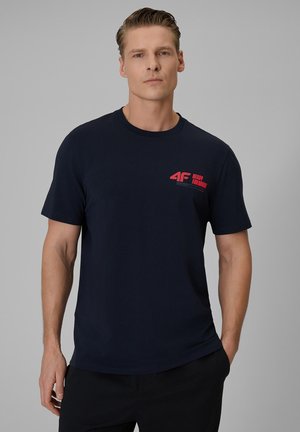 Mężczyzna ubrany w granatowy t-shirt z czerwonym logo "4F Ready For More" na lewej piersi, stojący na jednolitym, szarym tle.