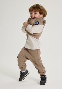 Beige vest med lynlås og et mærke, parret med en cremefarvet skjorte og beige joggers. Outfitten afsluttes med sorte sko med orange detaljer.