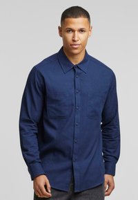 Camisa azul marino de botones con mangas largas, dos bolsillos frontales y cuello puntiagudo. Confeccionada con una tela suave de un solo color.
