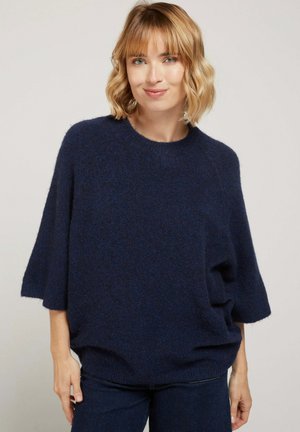 Femme aux cheveux blonds courts portant un pull ample bleu marine foncé et un pantalon assorti, debout devant un arrière-plan clair uni.