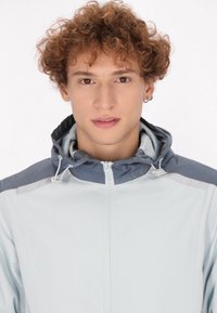 Giacca a vento azzurro chiaro con yoke blu scuro, dotata di chiusura frontale con zip e cordini sul cappuccio. Texture liscia.