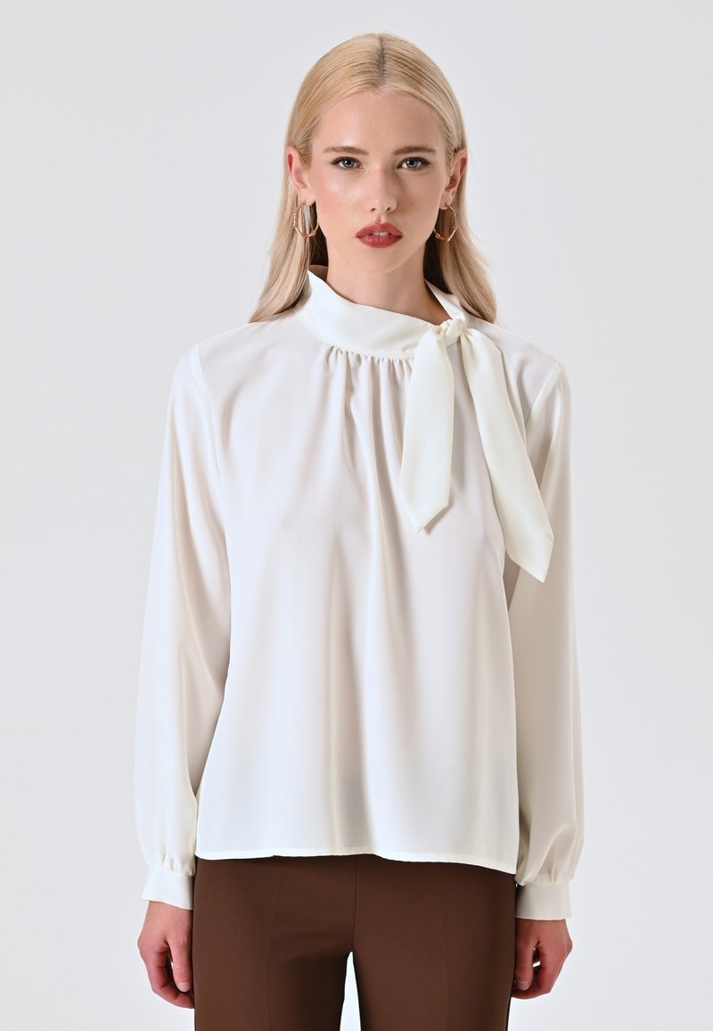Weißes Blusenshirt mit hohem Kragen, gerafften Details und einer gebundenen Schleifenakzent. Lange Ärmel mit Knopfmanschetten; glattes, leichtes Gewebe.
