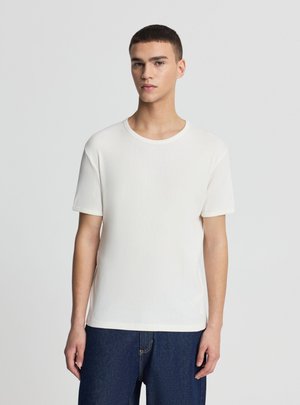 Giovane uomo con capelli corti e scuri che indossa una maglietta bianca semplice a maniche corte e jeans blu, in piedi davanti a uno sfondo grigio chiaro uniforme.