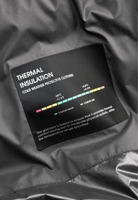 Etykieta izolacji termicznej na szarym materiale, wskazująca zakres komfortu od -24°C do +5,5°C dla odzieży ochronnej na zimne warunki.