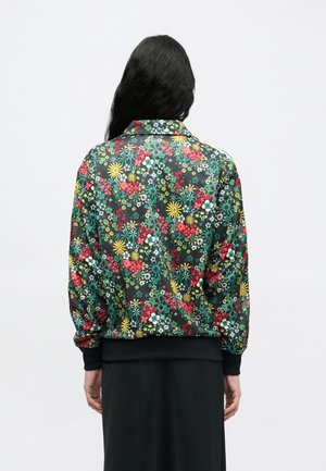Personne aux longs cheveux noirs portant une veste noire avec des motifs floraux colorés rouges, jaunes et verts, vue de dos.