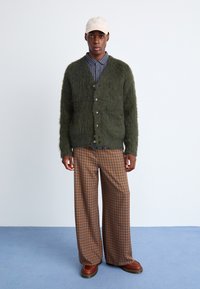 Πράσινο φουντωτό cardigan με κουμπιά, ψηλόμεσο καφέ καρό παντελόνι και μπεζ καπέλο. Το μοντέλο στέκεται σε μια ανοιχτή μπλε επιφάνεια μπροστά σε έναν απλό φόντο.