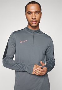Sivi Nike majica dugih rukava s patentnim zatvaračem, s ružičastim logotipom i crnim bočnim panelima. Glatka tekstura, prilagođen dizajn s otvorima za palčeve.