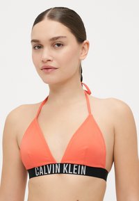 Koronkowy biustonosz w kolorze koralu o trójkątnym kształcie, z prążkowaną teksturą i cienkimi ramiączkami, z czarnym paskiem z logiem "CALVIN KLEIN" w białym napisie.